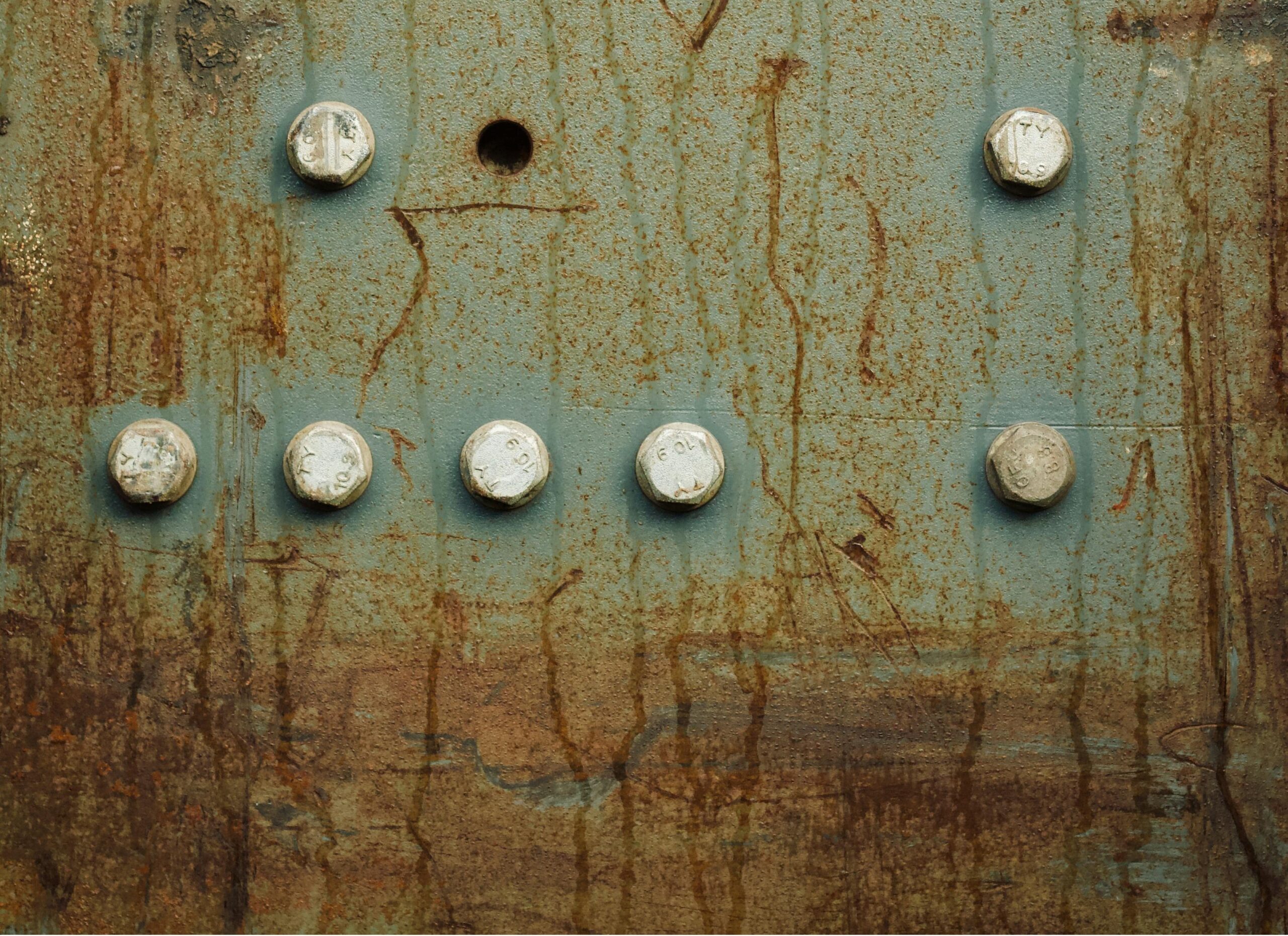 Close-up of a weathered teal metal panel with rust streaks and a row of bolts across the surface, creating a grid pattern.