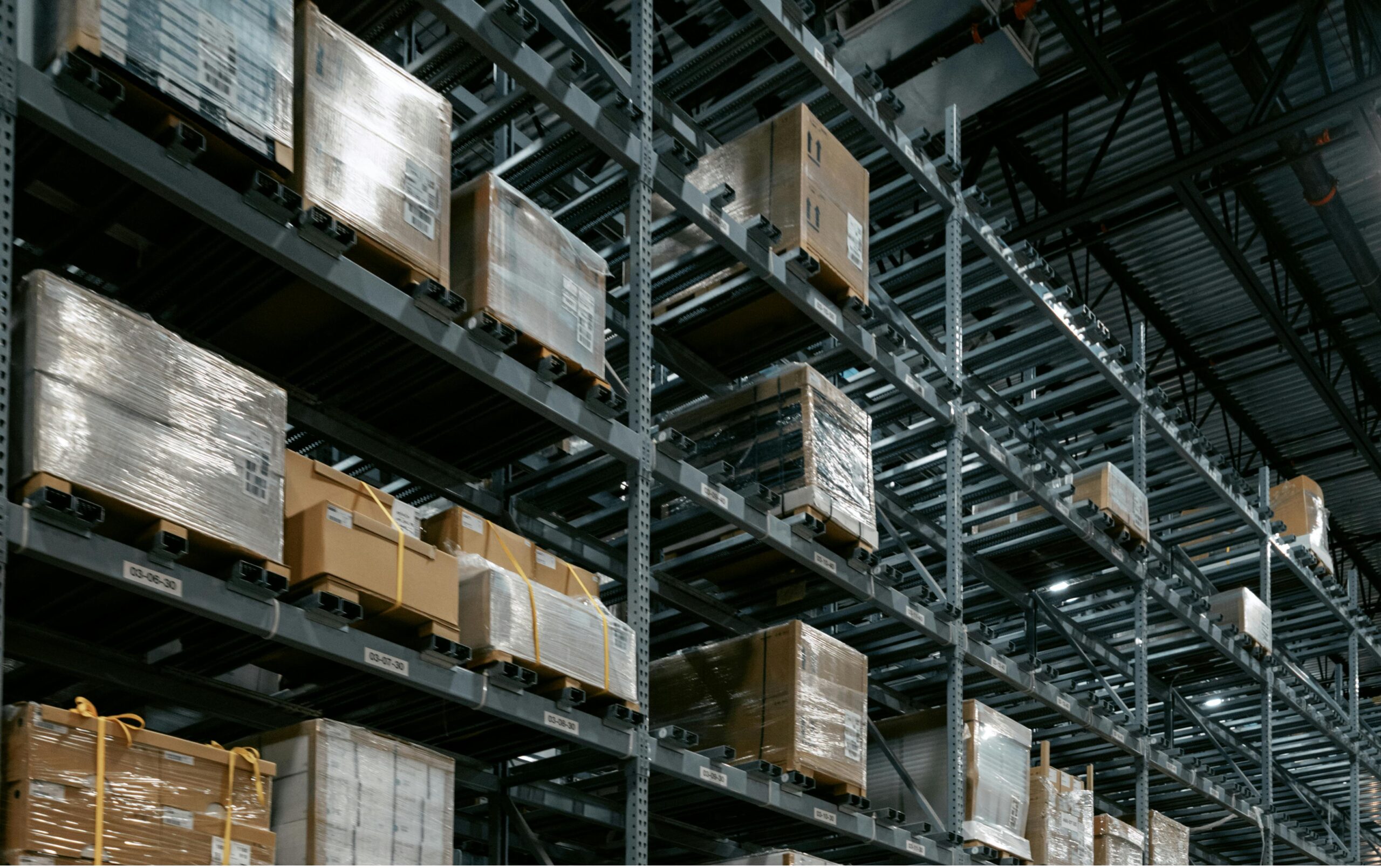Industrial warehouse with tall metal racks and palletized boxes wrapped in plastic on multiple levels