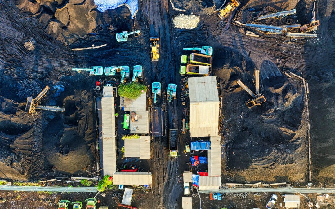 Aerial view of a construction site with blue and yellow heavy machinery, conveyors, and large dirt piles seen from above.