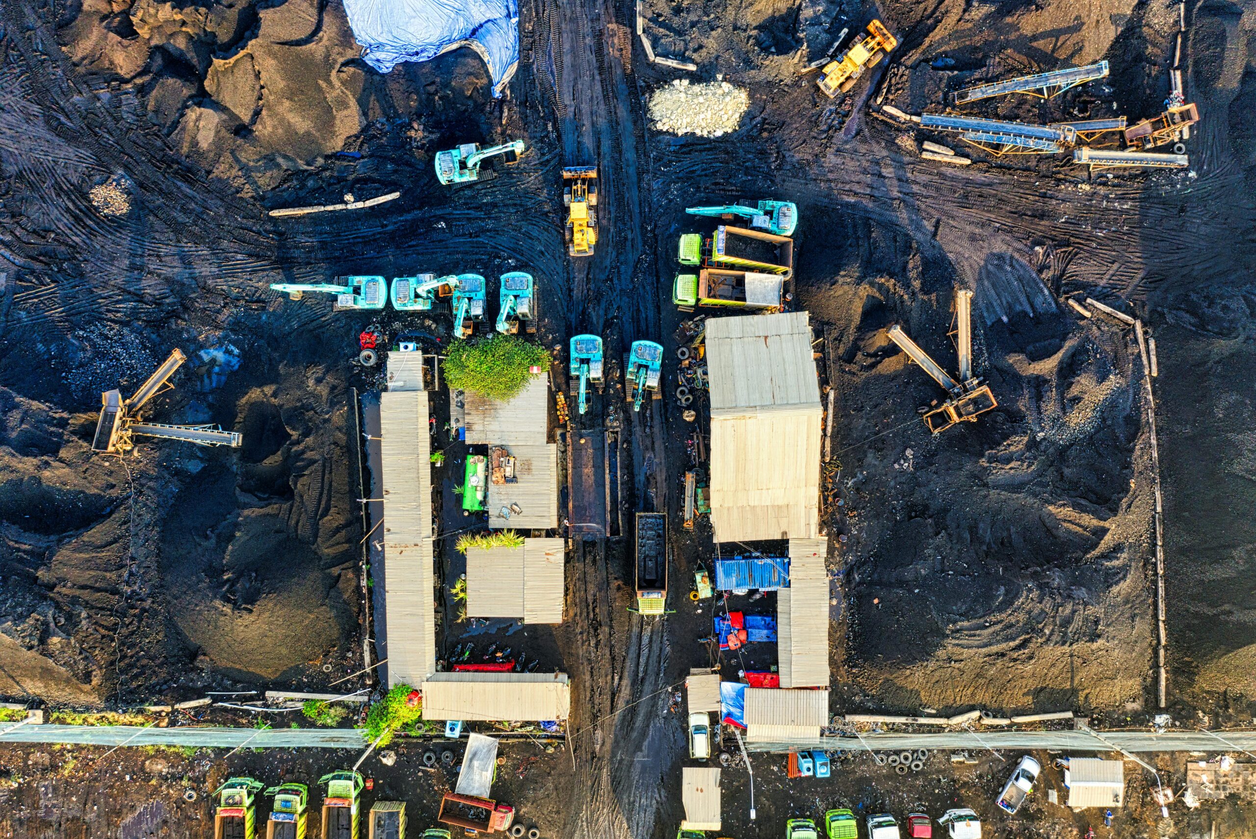 Aerial view of a construction site with blue and yellow heavy machinery, conveyors, and large dirt piles seen from above.